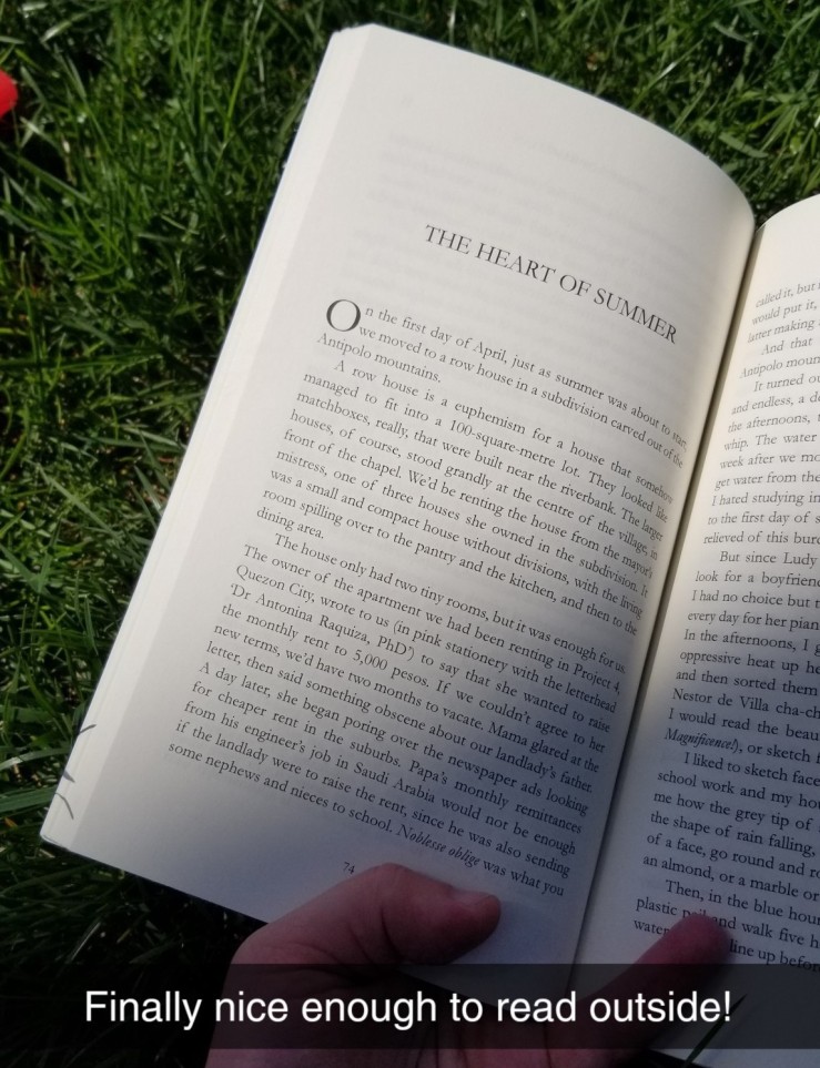 Image ID: The book "The Heart of Summer" held open against the grass. It is open to the titular short story, "The Heart of Summer." The title is in all caps, and the rest of the text on the page is not intended to be legible to read. The Snapchat caption reads "Finally nice enough to read outside!" End ID.